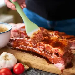 Ręka pędzlem smaruje żeberka marynatą. Obok miska z sosem, czosnek, pomidorki i papryczki chili. Ptaki zeberki problem w domu? Nie, to pyszne jedzenie!