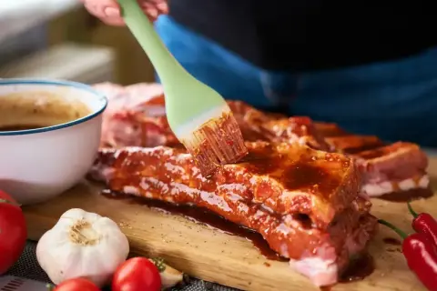 Ręka pędzlem smaruje żeberka marynatą. Obok miska z sosem, czosnek, pomidorki i papryczki chili. Ptaki zeberki problem w domu? Nie, to pyszne jedzenie!