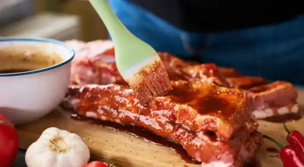 Ręka pędzlem smaruje żeberka marynatą. Obok miska z sosem, czosnek, pomidorki i papryczki chili. Ptaki zeberki problem w domu? Nie, to pyszne jedzenie!
