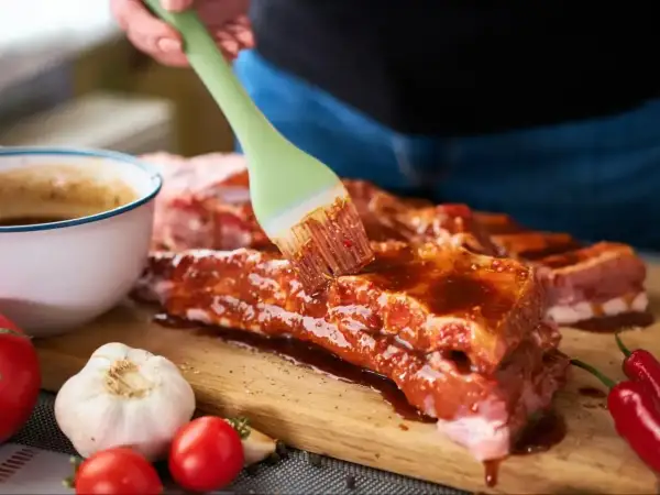 Ręka pędzlem smaruje żeberka marynatą. Obok miska z sosem, czosnek, pomidorki i papryczki chili. Ptaki zeberki problem w domu? Nie, to pyszne jedzenie!